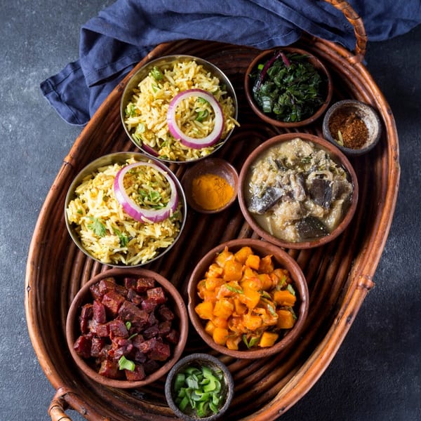 A woven tray holds various vegan dishes, including rice, curries, and vegetables in small bowls.