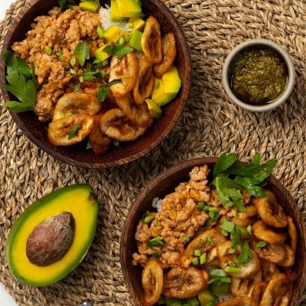 Two vegan bowls with plantains, avocado, and a side of green sauce on a woven mat.